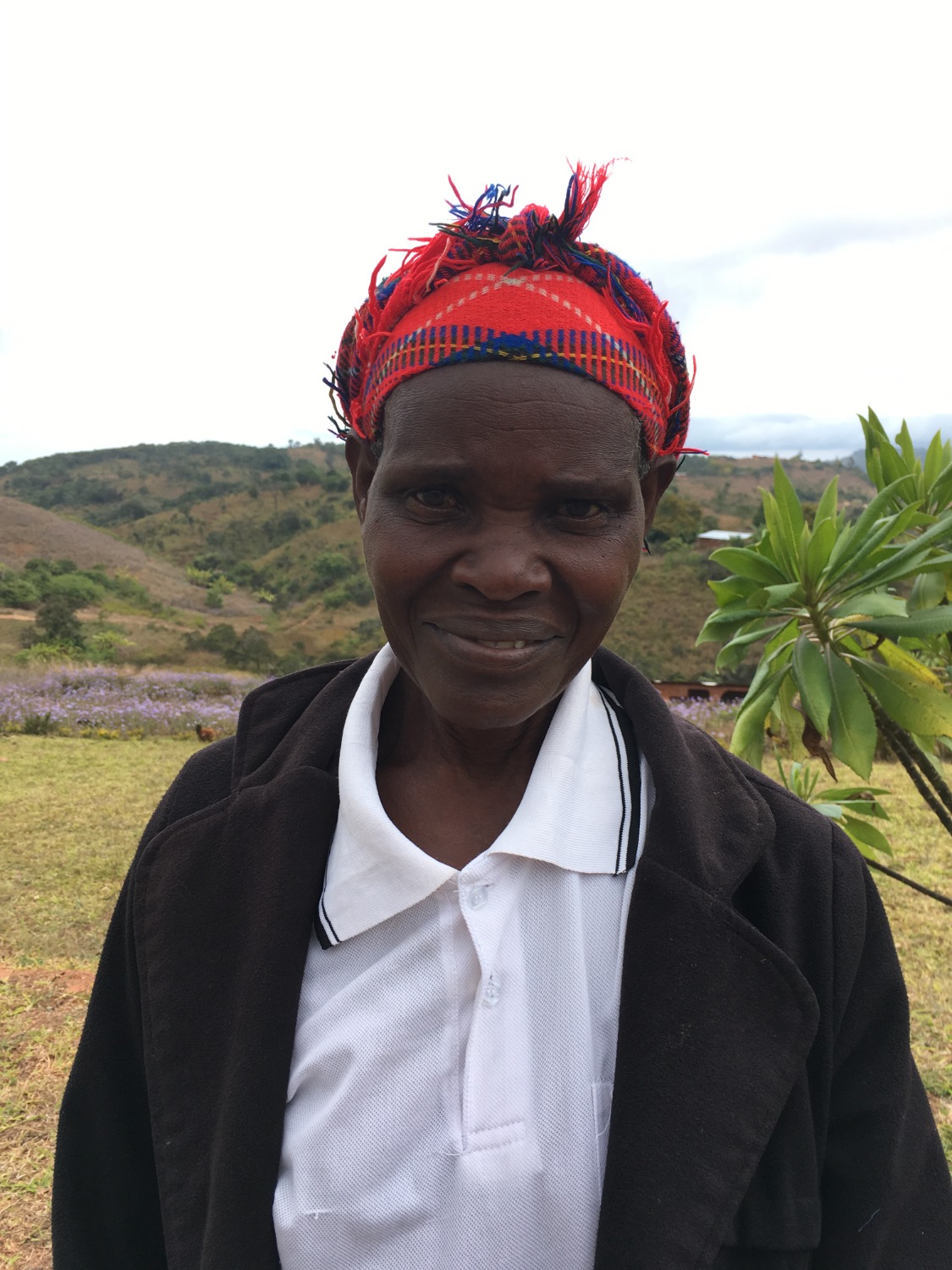 A woman in a red patterned headscarf and dark coat smiles gently, with rolling green hills and plants in the background.