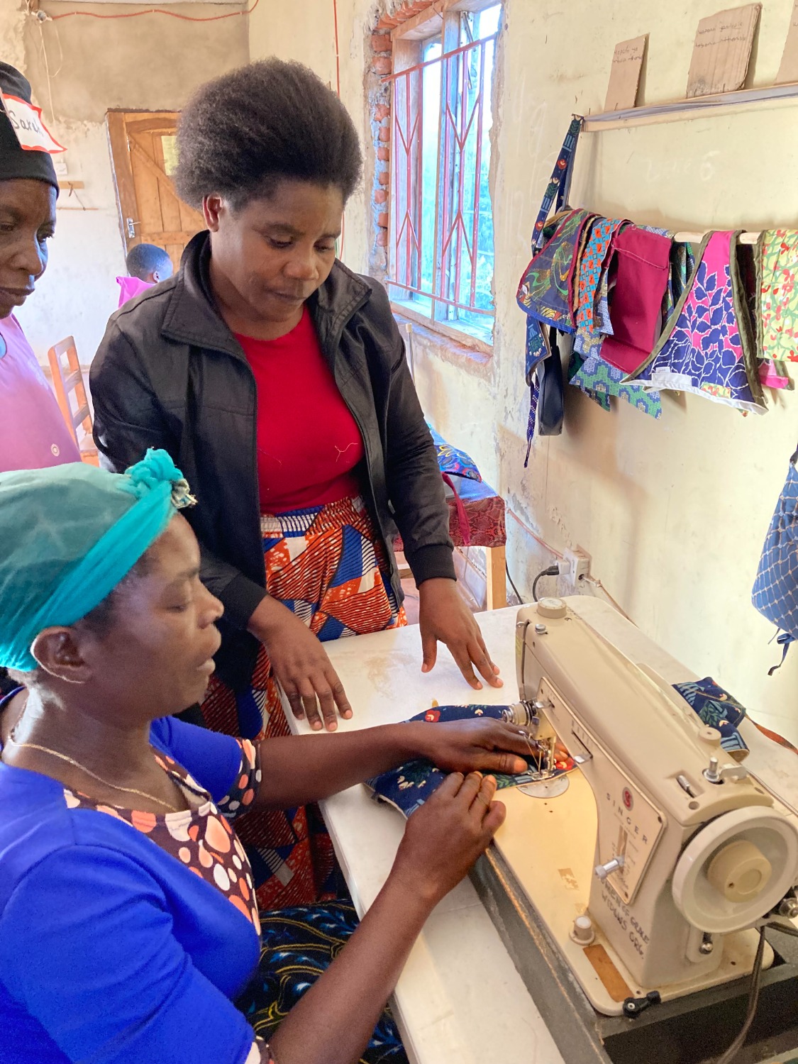 A woman in a blue top sews on a vintage Singer machine, guided by an instructor. Colorful fabric items hang on the wall.