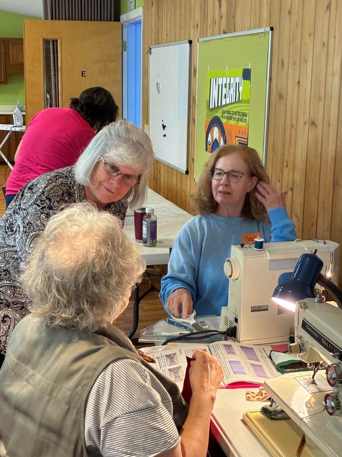 Three women gathered at a table for a sewing project. One works at a machine, another watches, while a third focuses on her craft.