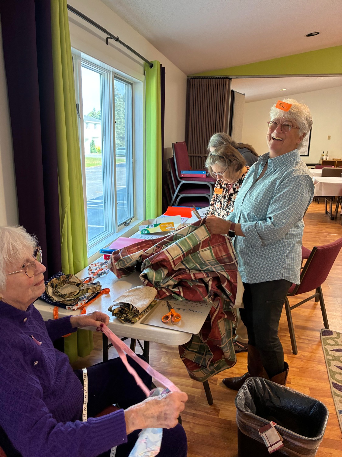 Two women in a brightly lit room. One laughs, holding plaid fabric. The other, seated, works with a pink ribbon. Craft items on table.